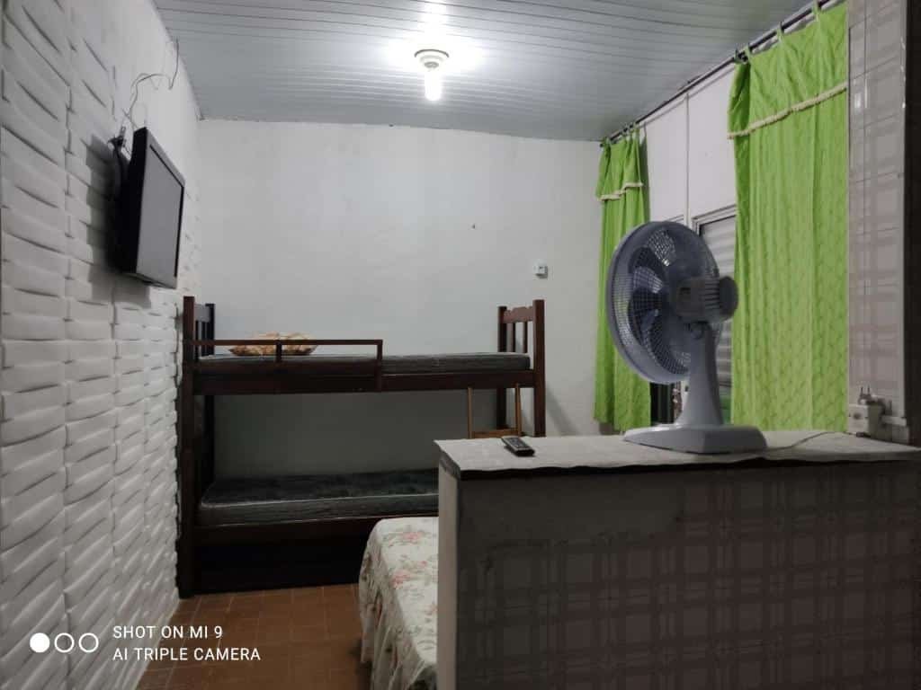 Pequeno quarto com beliche encostada na parede esquerda, tv centralizada na parede no meio do quarto, cama de casal ao lado de bancada com ventilador no lado direito do quarto. Imagem para ilustrar o post pousadas em Mongaguá.
