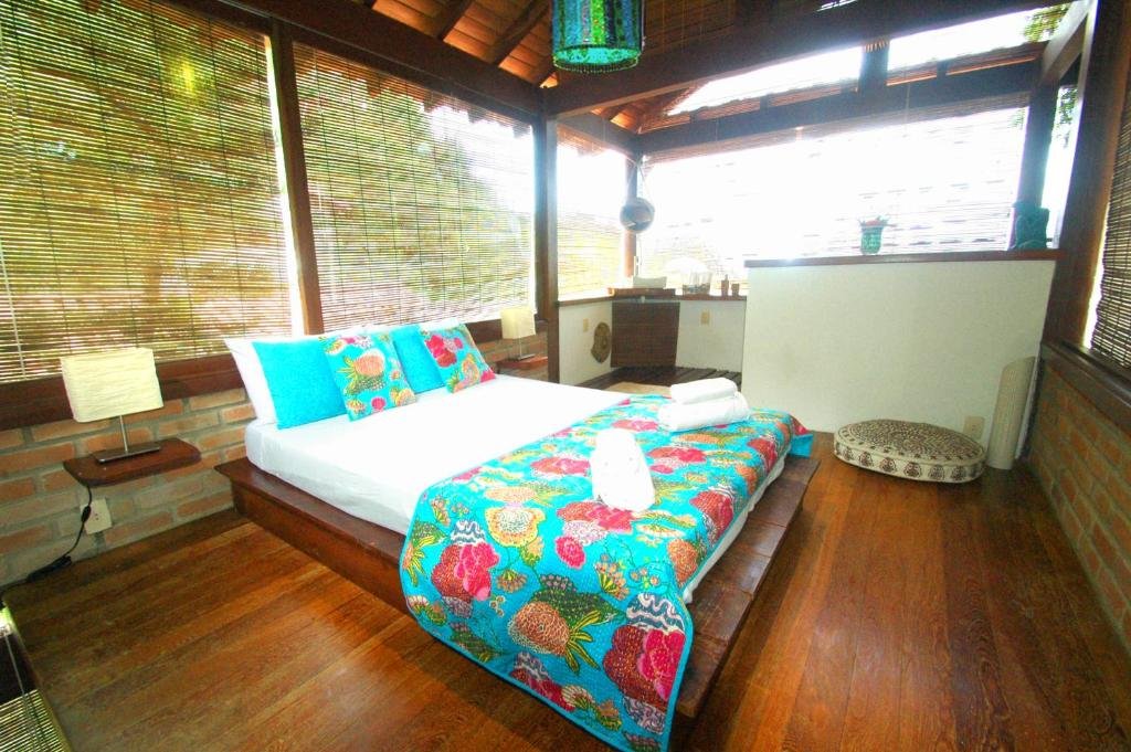Quarto da Casa Vista al Mar. Uma cama de casal do lado direito com uma prateleira e um abajur de cada lado. No fundo alguns utensílios do quarto. Em todas as paredes janelas de vidro e cortinas.