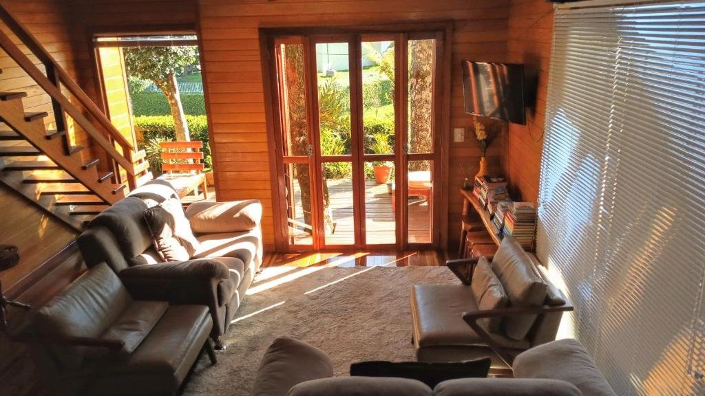 Uma sala de estar aconchegante na Casas Ana em Canela com paredes de madeira, janelas grandes, dois sofás, uma poltrona, uma TV, livros e vista para um jardim ensolarado.