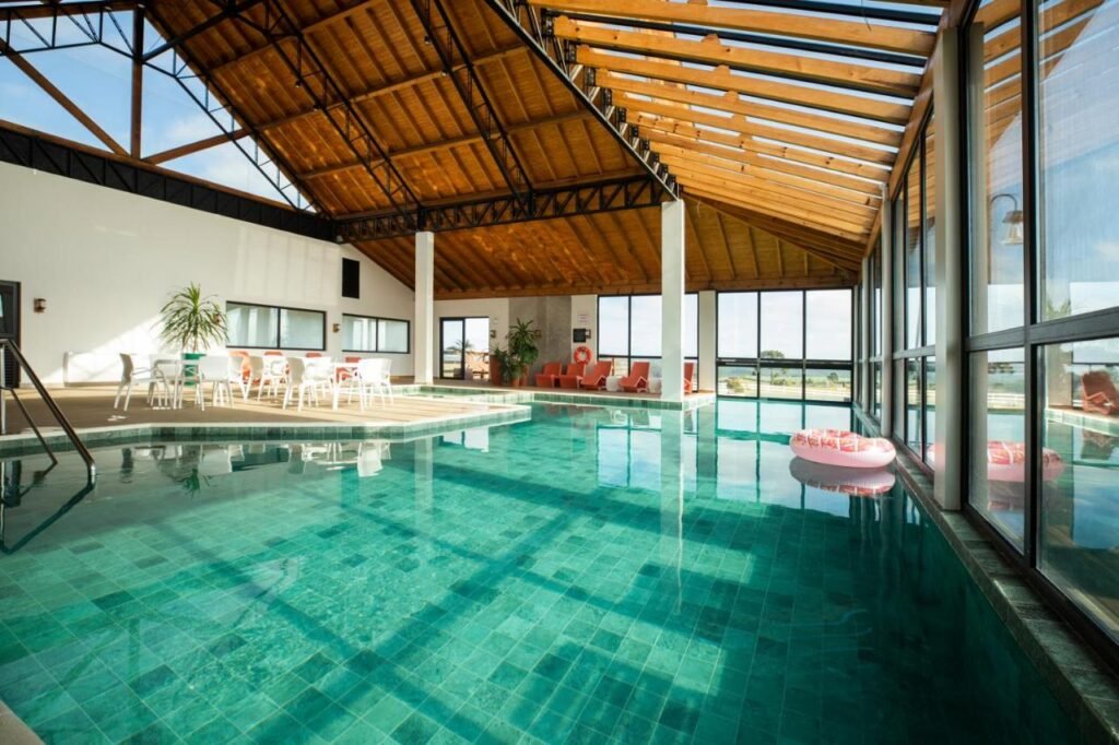 Piscina coberta com água limpa, cercada por cadeiras, mesas e plantas em vasos. Um teto de madeira e grandes janelas são visíveis. Esse é o Cerro Azul Hotel Fazenda, um dos hotéis fazenda em Santa Catarina.