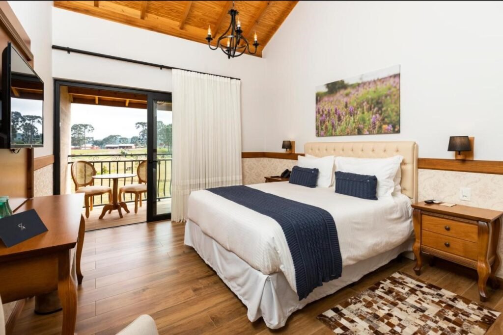 Um quarto aconchegante do Cerro Azul Hotel Fazenda com cama de casal, mesas de cabeceira, TV, varanda com vista e detalhes em madeira no teto e no piso.