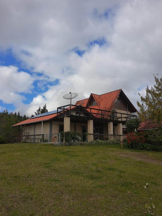 Uma casa de dois andares com telhado de telhas vermelhas, uma antena parabólica na varanda e um grande quintal gramado sob um céu nublado na Chácara Quedinha d