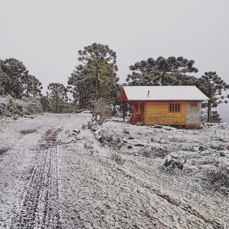 A imagem mostra a Casa de Campo Santa Paulina, um dos chalés na Serra Catarinense, uma pequena casa de campo construída com madeira, localizada em uma paisagem de inverno com o solo e árvores cobertos por neve fina. A casa tem um telhado branco, com detalhes rústicos, situada em meio a árvores típicas da serra, como araucárias. O cenário transmite uma sensação de isolamento e tranquilidade, com a vegetação ao redor suavemente coberta pela neve, criando um ambiente aconchegante e sereno.