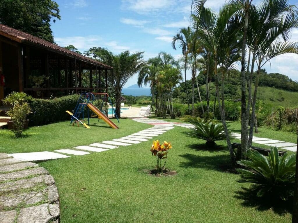 Área de lazer do Chalé Boiçucanga. Um pequeno parquinho está em um gramado, cercado por algumas árvores e paisangem cheia de vegetação. Ao fundo é possível ver uma piscina.