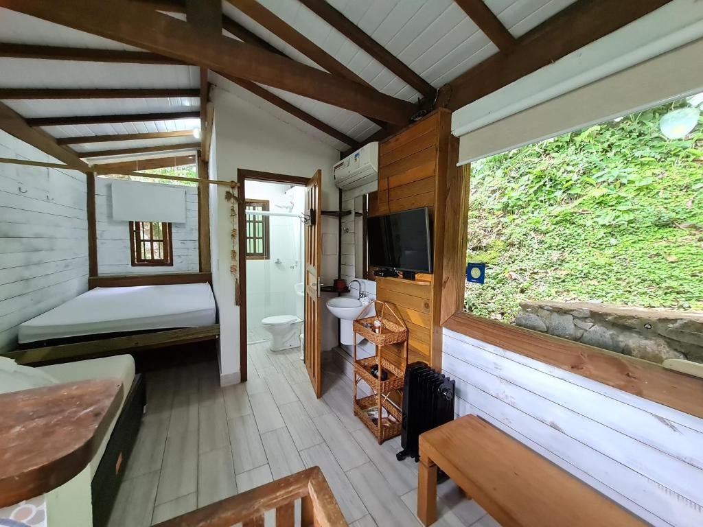Foto do quarto no Residencial Marina Del Sol. O interior do chalé é de madeira. Na direita há uma janela, uma TV, ar-condicionado e pia. No fundo à esquerda há uma cama de casal. No centro há uma porta para o banheiro.