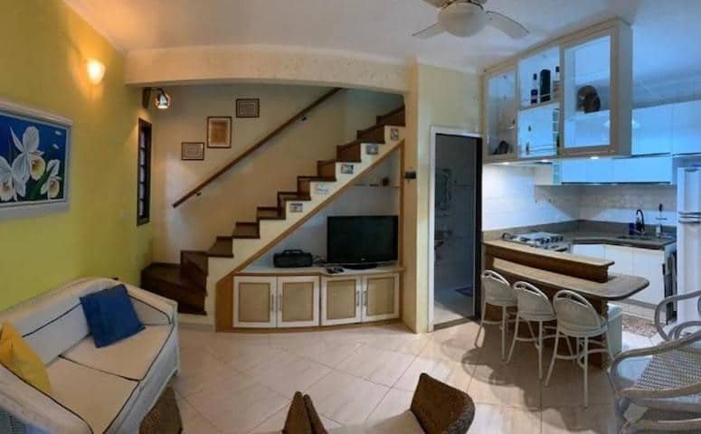 Sala e cozinha do Chalé Recanto do Barão. Do lado direito, na sala, um sofá e uma poltrona ficam do lado oposto da tv, que está embaixo da escada. Já do lado esquerdo há um balcão com algumas cadeiras separando o ambiente com uma pia, fogão e geladeira.