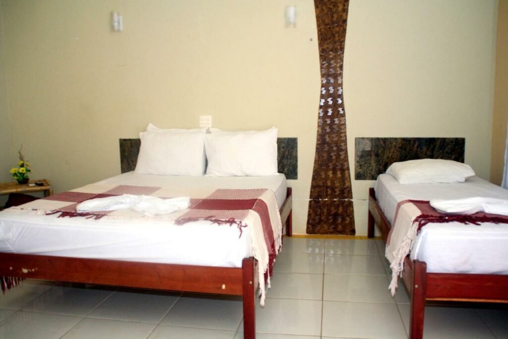 Quarto da Chalemar Hotel Pousada. Uma cama de casal está do lado esquerdo e uma cama de solteiro está do lado direito. No canto da imagem, ao lado da cama de casal, há uma pequena mesinha. Esta é mais uma das pousadas em Baía Formosa, RN.