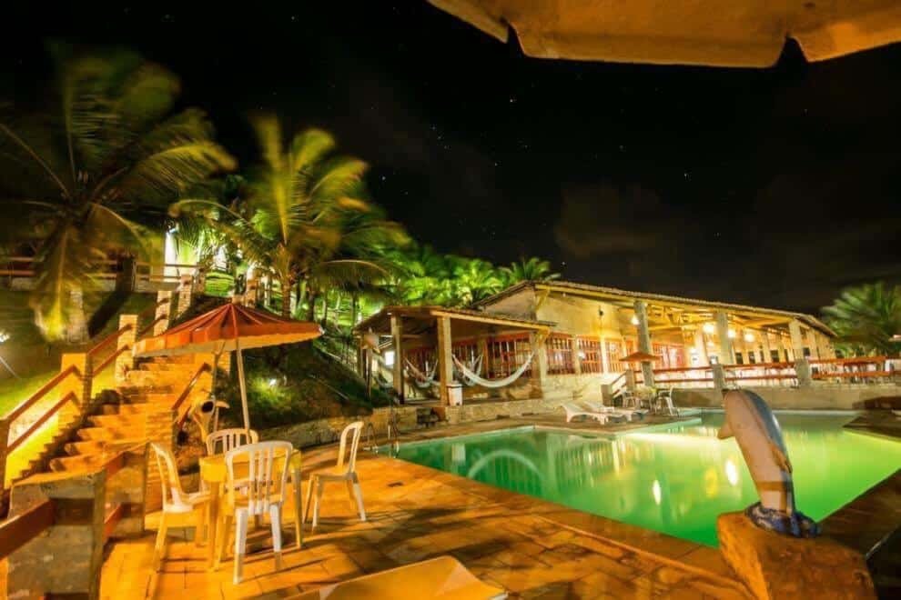 Área da piscina da pousada Chalemar Hotel Pousada. A piscina é grande, está no meio e é rodeada por espreguiçadeiras, mesas e cadeiras e guarda-sóis. No canto da imagem há uma escada levando par a parte de cima da pousada.