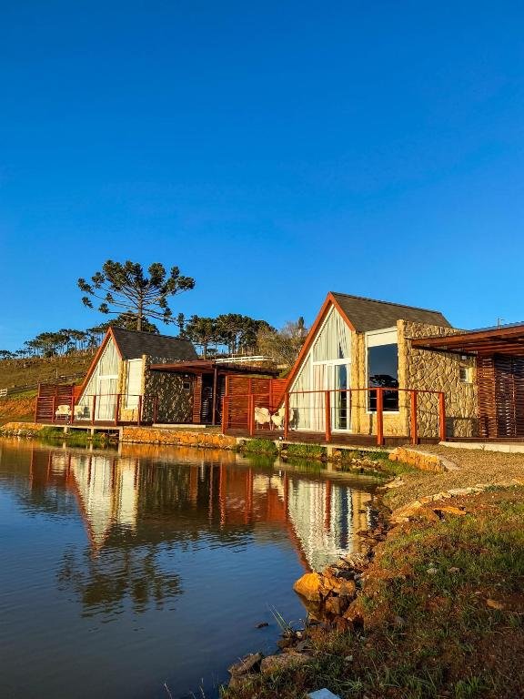 A imagem mostra os chalés na Serra Catarinense do Recanto Entre Rios dispostos em fila, à beira de um lago sereno, refletindo perfeitamente suas formas na água. Os chalés possuem uma arquitetura em estilo "A-frame", com paredes de pedra e grandes janelas de vidro que capturam a luz do sol da tarde. Cada chalé tem um deck de madeira com guarda-corpo, proporcionando um espaço privado ao ar livre com vista para o lago. Ao fundo, as araucárias típicas da serra se destacam contra o céu azul claro, completando a paisagem natural tranquila e acolhedora.
