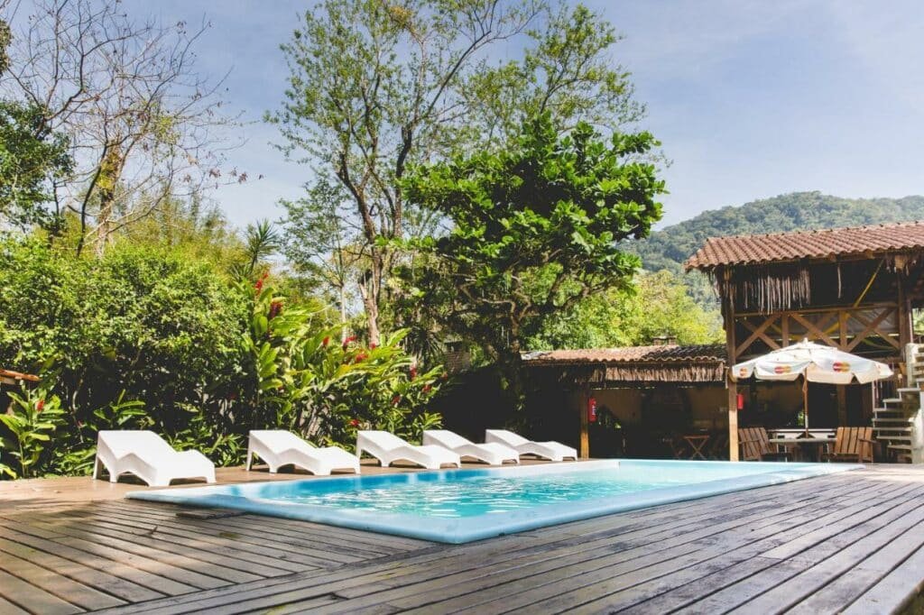Piscina do Chalés Água de Coco. Ela está em um chão de madeira, ao lado de várias espreguiçadeiras. Ao fundo estáo algumas áreas do chalé.
