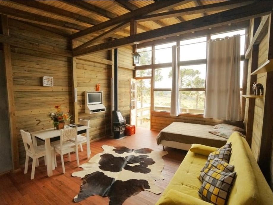 Um interior rústico de madeira com uma cama, sofá amarelo, pequena mesa de jantar, TV montada na parede e grandes janelas com vista para a vegetação nos Chalés da Serra Catarinense, um dos hotéis fazenda em Santa Catarina.