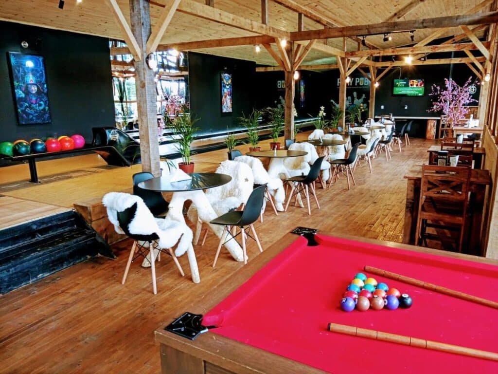Um espaço recreativo com mesa de sinuca, pistas de boliche e área de estar com mesas e cadeiras cobertas de pele branca sobre piso de madeira nos Chalés da Serra Catarinense.