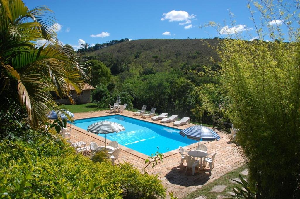 A imagem mostra uma piscina retangular cercada por espreguiçadeiras brancas e mesas com guarda-sóis listrados de azul e branco. Ao redor da piscina, há uma área pavimentada e muita vegetação verde, com plantas tropicais e árvores ao fundo. Além disso, o cenário é composto por colinas verdes sob um céu azul claro com poucas nuvens. O ambiente parece ser uma propriedade rural ou pousada em uma área montanhosa, transmitindo tranquilidade e contato com a natureza. Esse é o Chalés de Minas Hotel Fazenda, um dos hotéis-fazenda em Minas Gerais.