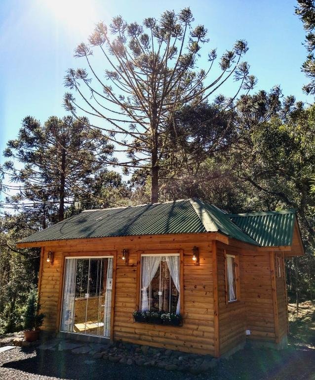 Fachada do Chalés Estância Cachoeira do Avencal em Urubici em estilo de madeira com janela e portas de vidro em que é possível observar a natureza, há ainda diversas araucárias de fundo.