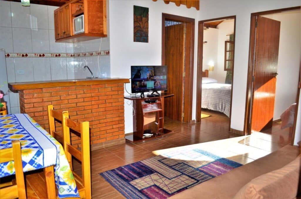 Uma pequena sala de estar nos Chalés Fazenda Vale da Mata com TV, balcão de cozinha de tijolos, cadeiras de madeira e uma toalha de mesa colorida. Um quarto é visível através de uma porta aberta.