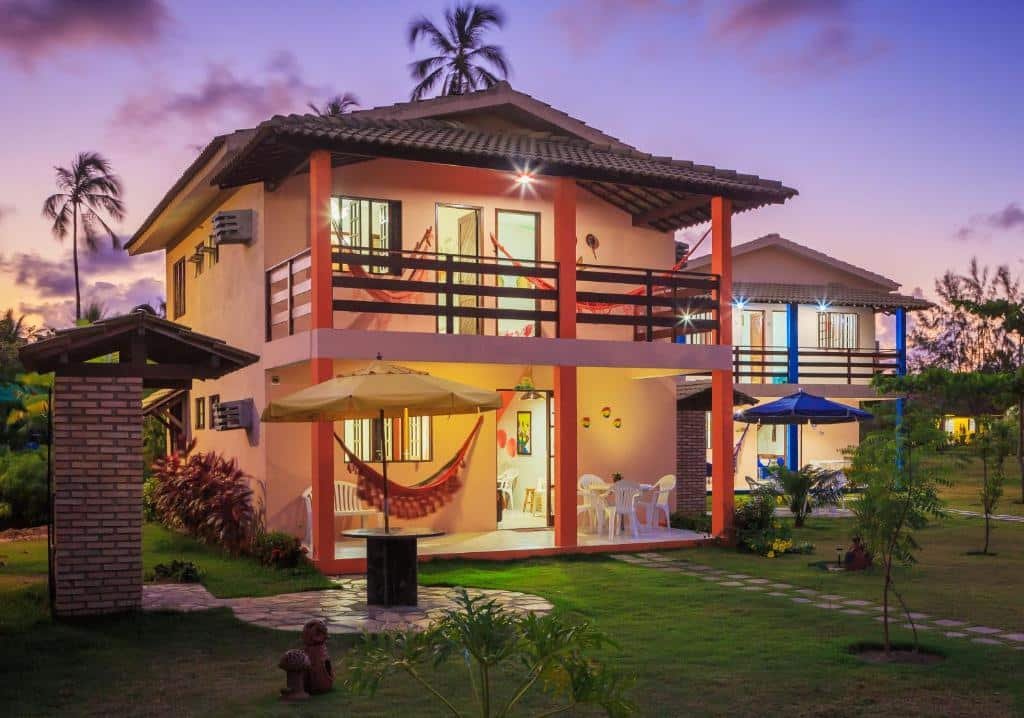 Uma das casas do Chales Maragogi, sendo o local um osbrado com luzes acesas durante um inicio de noite. O local tem uma varanda no térreo com redes, banco e mesa de plástico com cadeiras e uma porta dando acesso ao interior. No andar de cima tem uma sacada com duas redes e duas portas abertas. À direita tem uma casa semelhante, e o local é cercado de gramado e árvores.