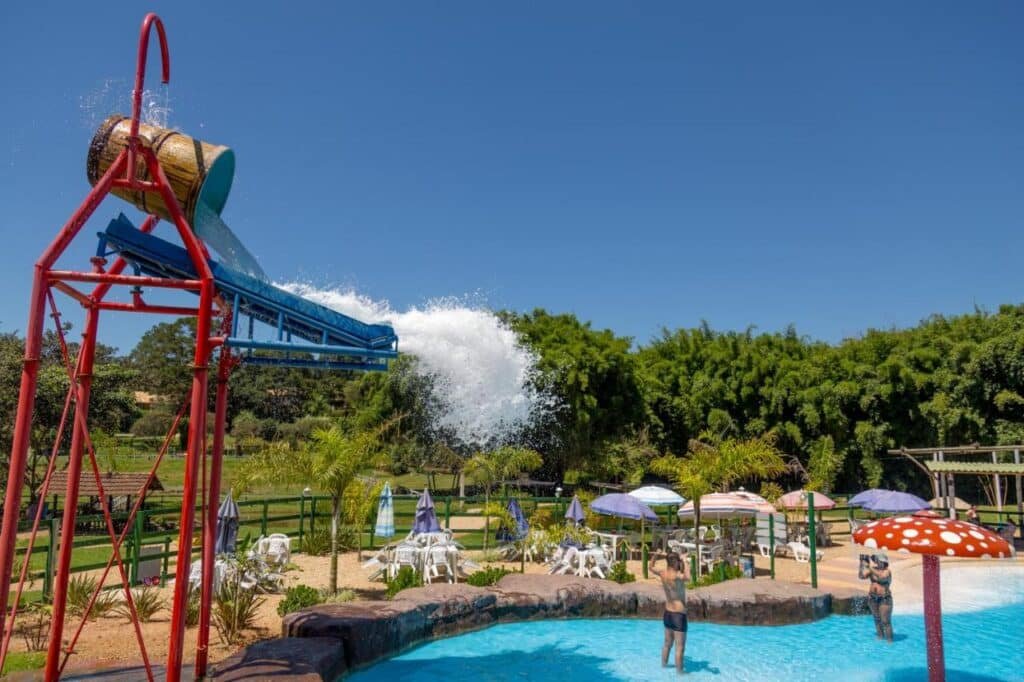 A imagem mostra uma piscina infantil em um parque aquático dos Chalés Praia de Minas, com um grande balde suspenso por uma estrutura vermelha. O balde está despejando uma quantidade significativa de água em um escorregador azul, criando uma cascata para os banhistas abaixo. Na piscina, há um cogumelo vermelho com bolinhas brancas, que serve como um chafariz Esse local é um dos hotéis-fazenda em Minas Gerais.de água. Algumas pessoas estão na piscina, brincando e se refrescando, enquanto ao fundo, mesas e cadeiras com guarda-sóis coloridos estão dispostas em uma área de descanso cercada por vegetação verde. O céu está claro e azul.