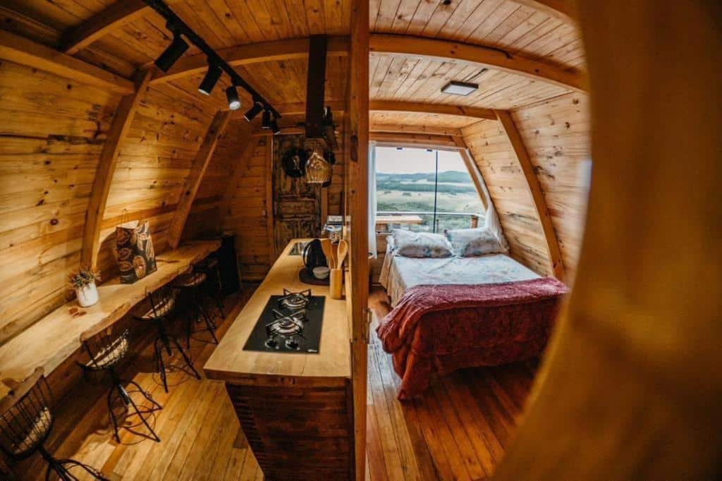 O interior de uma cabana de madeira com uma pequena cozinha e uma cama perto de uma grande janela com vista para uma paisagem rural nos Chalés Recanto do Vale, um dos hotéis fazenda em Santa Catarina.