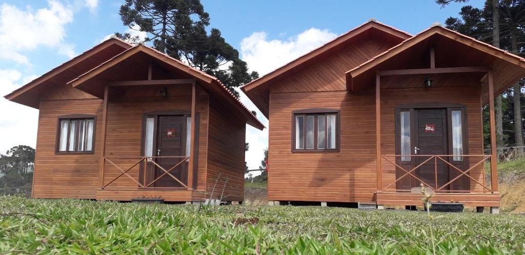 fachada do Chalés Sossego da Serra sendo casinhas de madeira com porta e janela de vidro, há uma grama baixa bem cuidada abaixo, o céu é azul com nuvens e há araucárias atrás