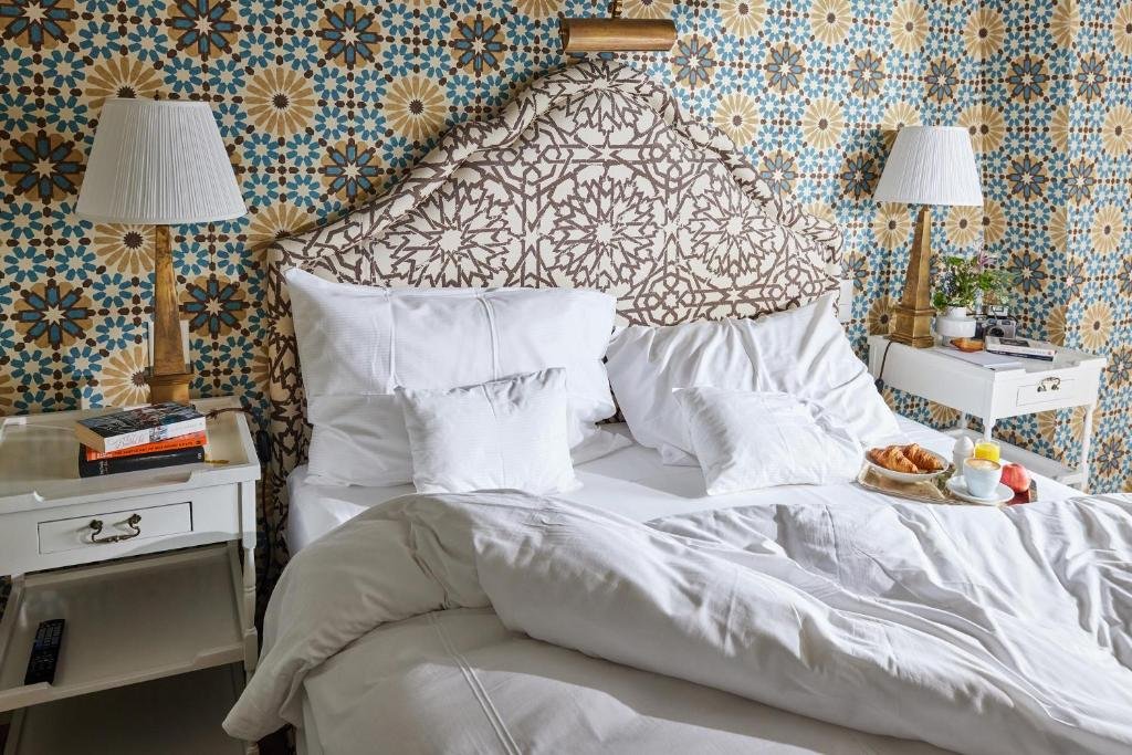 Foto do Château Gütsch, para representar o post de hotéis em Lucerna. Temos uma visão de perto da cama, que é de casal e com lençóis brancos. Em cima dela também há uma bandeja com café da manhã. Em ambos seus lados existe uma mesa de cabeceira branca com abajur, livros e vasos de planta em cima. A parede atrás possui um papel de parede de flores nas cores branco, azul e amarelo.