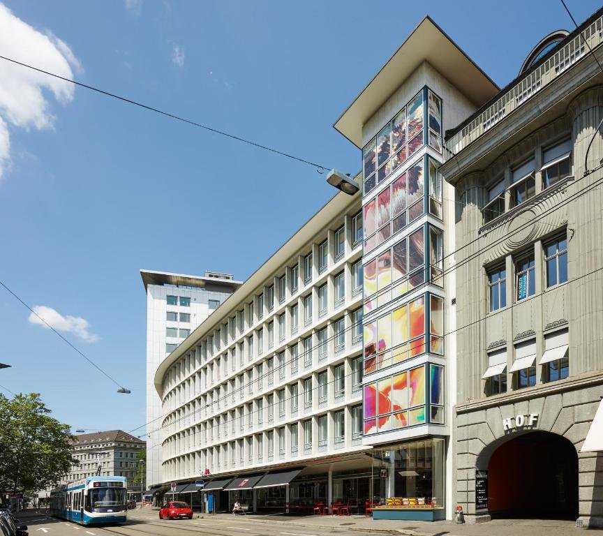 Prédio do citizenM Zürich num dia ensolarado, com céu azul, com várias janelas e rua com trilho de metrô bem a frente