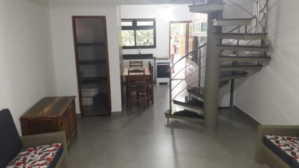 Espaço interno do Condominio Frente Praia Boissucanga. Ao fundo é possível ver a cozinha e uma mesa perto de uma escada. Em primeiro plano estão dois sofás um de frente para o outro e um baú encostado na parede do lado esquerdo.