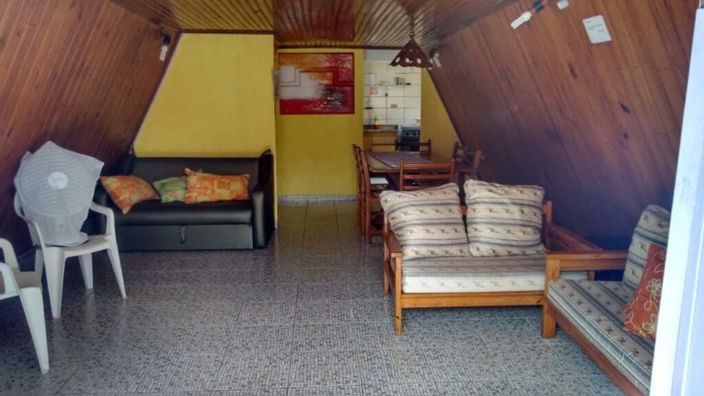 Condomínio Sol Mar e Cia. Bem ao fundo é possível ver o pedaço de uma cozinha e logo em seguida uma mesa de seis lugares. Mais a frente três sofá estão compondo o ambiente, que possui o teto e duas paredes de madeira.