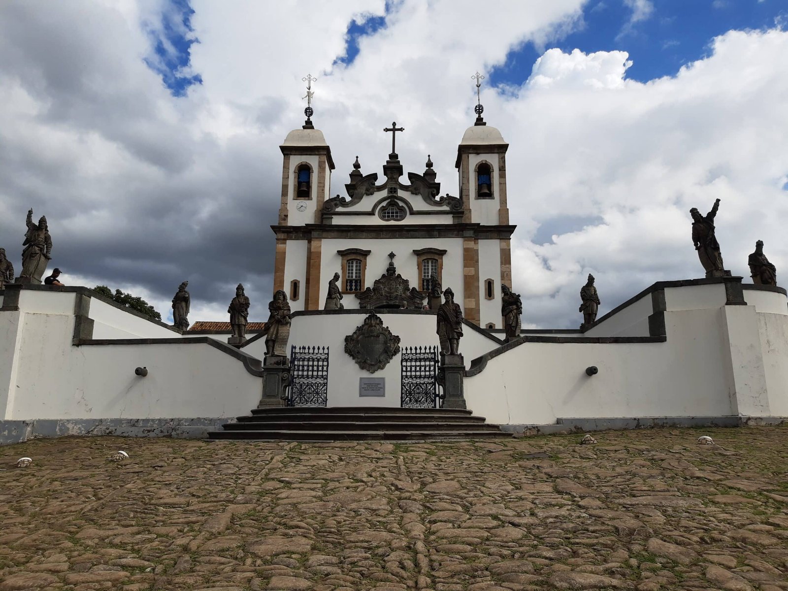 vista frontal externa do Santuário do Bom Jesus de Matosinhos onde é possível enxergar todas as doze estátuas esculpidas por Aleijadinho. Além disso, há também a igreja histórica do século 18 mais ao fundo.