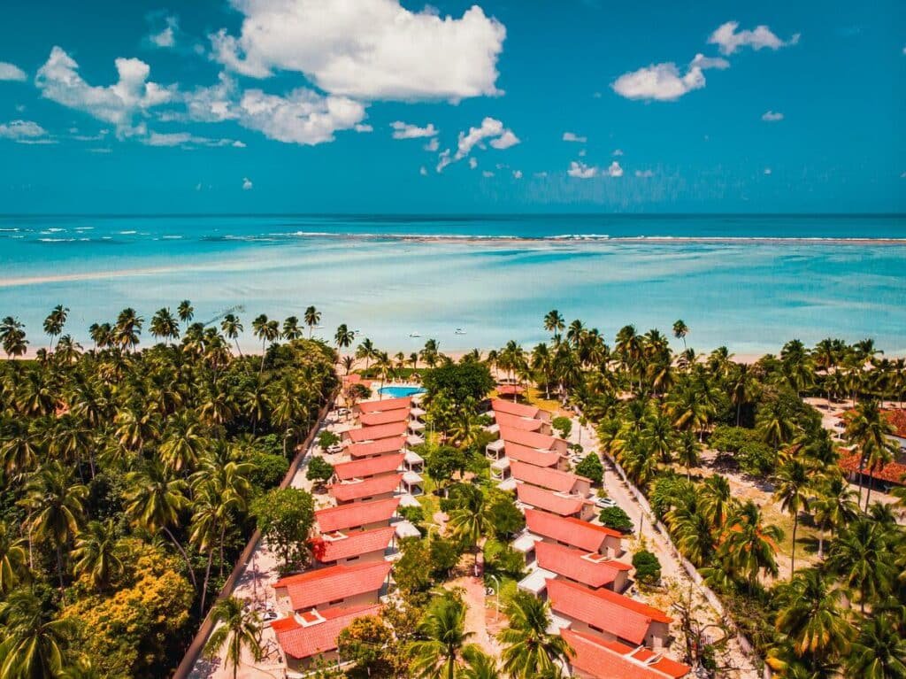 Vista aérea do Costa Dourada Village, um dos resorts em Maragogi. Na imagem, há duas fileiras de bangalôs com tetos no formato de triangular. Nos lados há árvores e coqueiros e, à frente, um mar azul turquesa