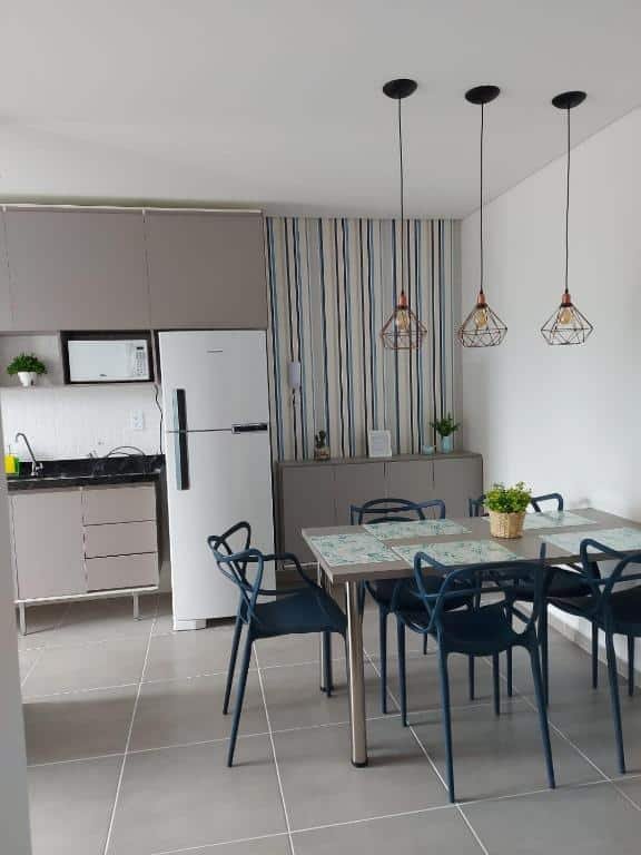 Sala de estar e cozinha no Apartamento Completo em Ubatuba - Tenório - Piscina e Churrasqueira gourmet, tudo decorado e novo, uma mesa com cinco lugares, uma geladeira, três pequenos lustres e armários, para representar pousadas na Praia do Tenório