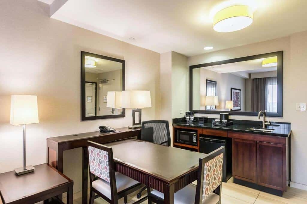 Imagem de uma cozinha no Embassy Suites by Hilton Newark Airport. A cozinha é equipada com armários brancos e bancada de granito. Na parte superior, há armários suspensos com pratos, copos e utensílios de cozinha organizados. Na bancada, há uma pia com torneira, um fogão com quatro bocas e um forno embutido. Há também uma geladeira, um micro-ondas e uma cafeteira. A cozinha oferece um espaço funcional para preparar refeições e possui todos os utensílios necessários. O Embassy Suites by Hilton Newark Airport é um dos hotéis perto do aeroporto de Newark.