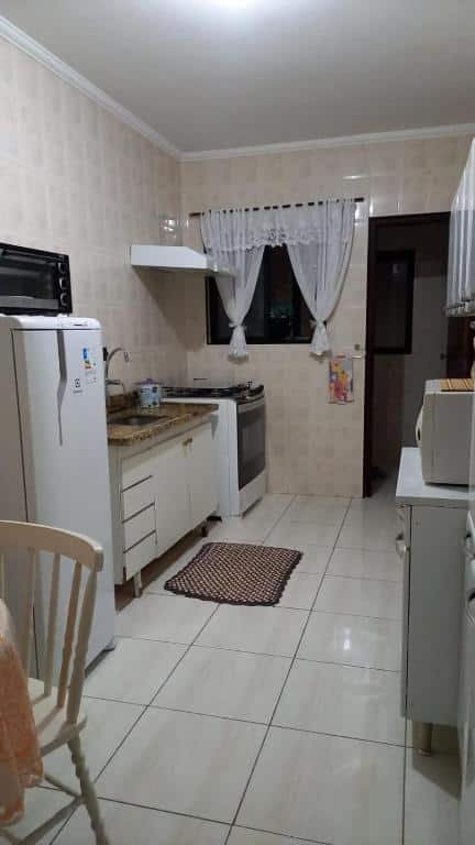 Cozinha com pequena janela com cortina e porta na parede aos fundos, fogão, pia e geladeira no lado esquerdo e armários com microondas no lado direito.