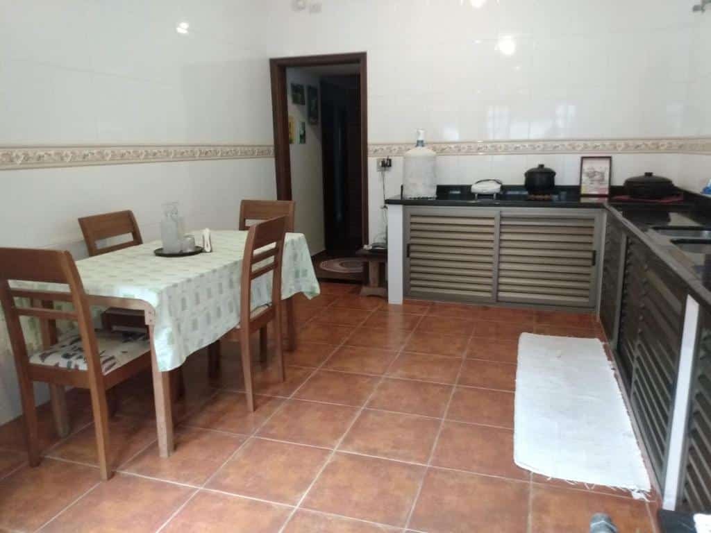 Cozinha da Hospedaria Namastê. Uma mesa de madeira com quatro cadeiras no lado esquerdo. No lado direito, a pia e os armários em baixo da pia. No lado esquerdo, no fundo, a porta da cozinha.