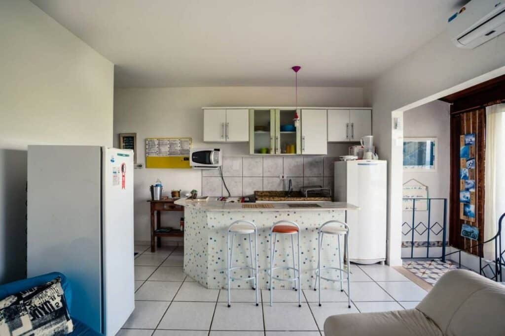 Cozinha do Loft com vista ao mar com geladeira do lado esquerdo da imagem, do lado direito banquetas e um balcão e ao fundo do balcão armário e outra geladeira.