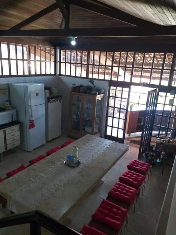 Cozinha do Sefiroh Hostel. No meio há uma mesa grande com dois bancos grandes em cada lado da mesa. No lado esquerdo há duas geladeiras, um armário com portas de vidro e é possível ver um pouco de outro armário com um microondas em cima. No lado direito há uma porta de madeira com detalhes em vidro aberta.