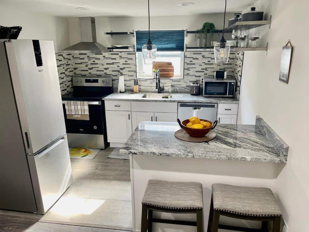 cozinha do Cozy 1BR apt in Elizabeth com uma geladeira de inox no lado esquerdo, um balcão de pedra com dois bancos estofados e sem encosto no lado direito, e uma bancada de pedra ao fundo, com uma pia, microondas de inox, fogão de inox, sugar, duas prateleiras e uma janela acima da pia.