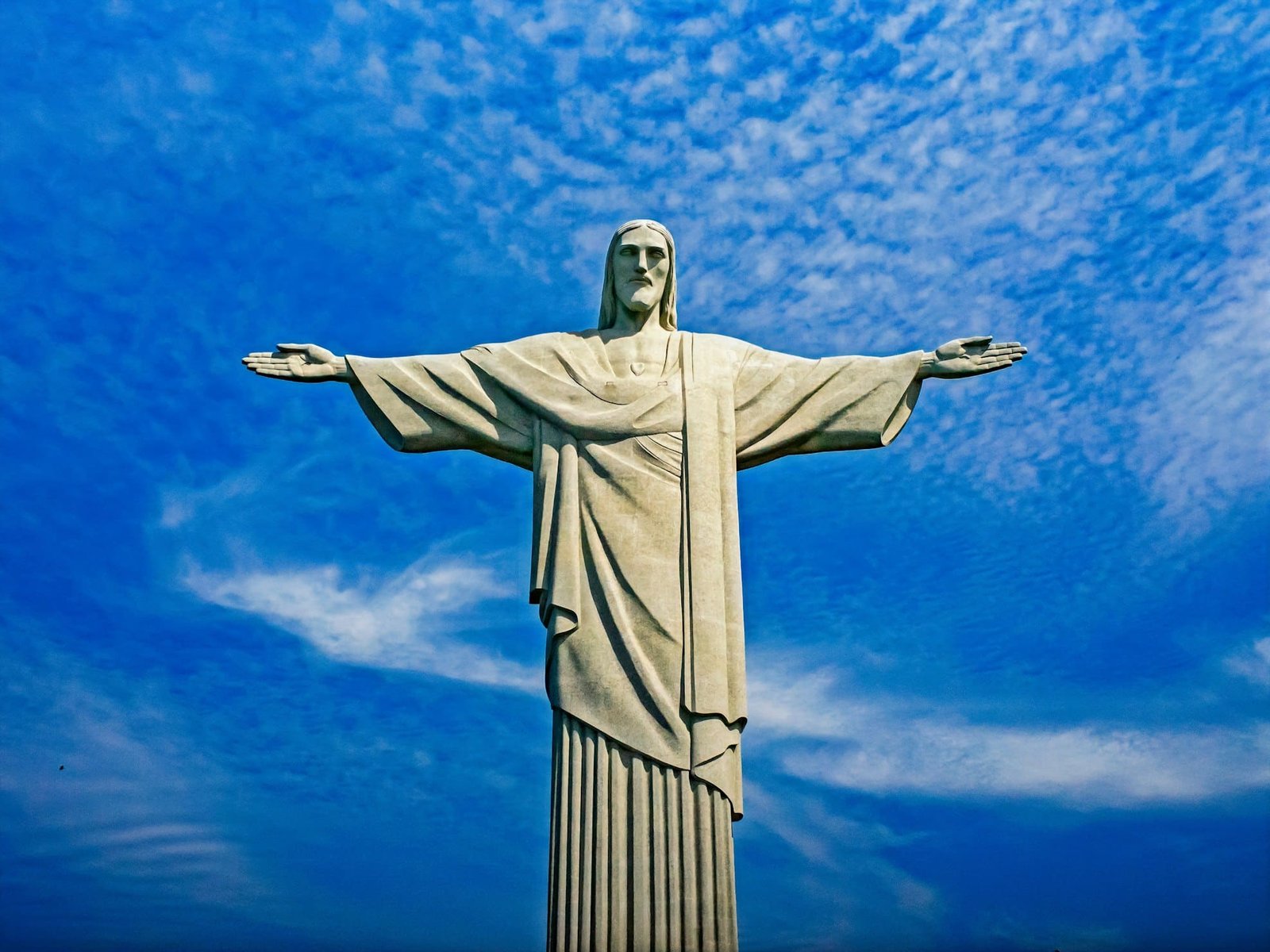 Estátua do Cristo Redentor com os braços estendidos, em contraste com um céu azul vibrante com nuvens dispersas.
