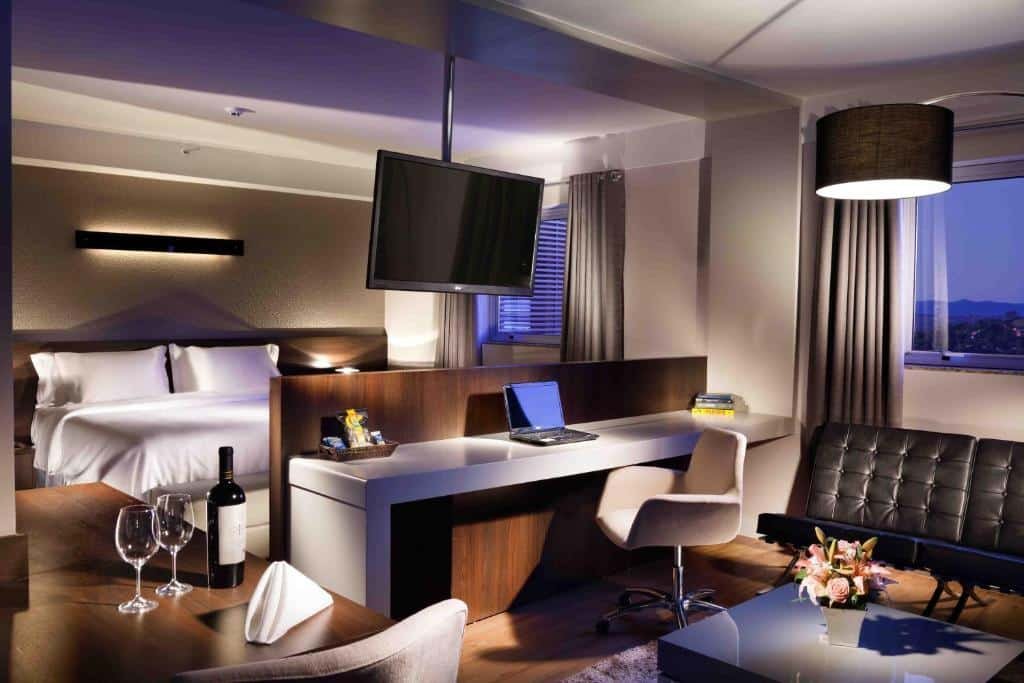 Quarto do Delmond Hotel com uma cama de casal, uma mesa com cadeira e notebook, televisão e outra mesa com vinho e duas taças. É possível ver também janelas, um sofá e uma mesinha pequena com um vaso de flor.