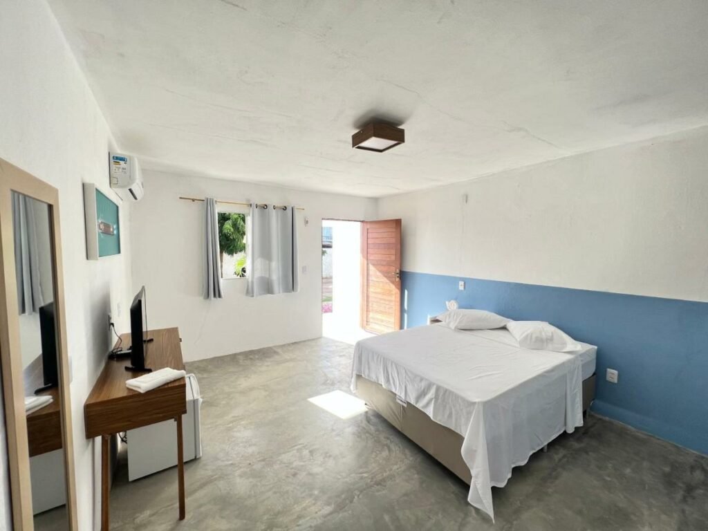 Quarto da Dlux Suites Baia Formosa. Ele é bem espaçoso e conta com uma cama de casal bem no meio, de frente para uma tv em cima de uma mesa, um mini frigobar e um espelho. Do lado esquerdo estão a janela e a porta.