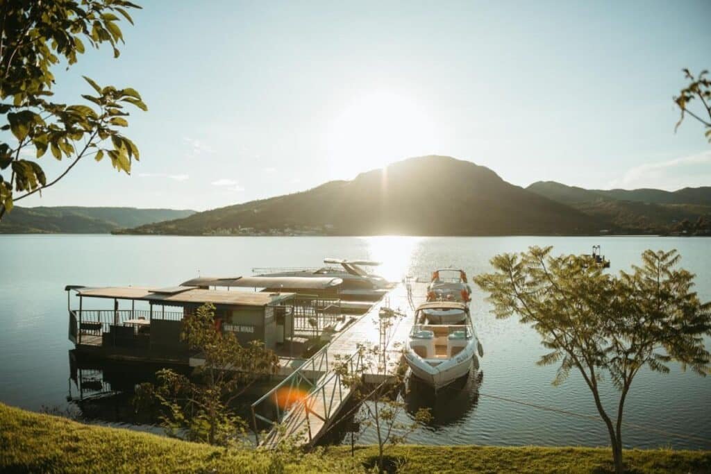 Barcos atracados em um pequeno píer em um lago calmo, cercado por árvores, com uma montanha e o pôr do sol ao fundo. Esse local fica no Engenho da Serra Hotel EcoResort, um dos hotéis fazenda em Minas Gerais.
