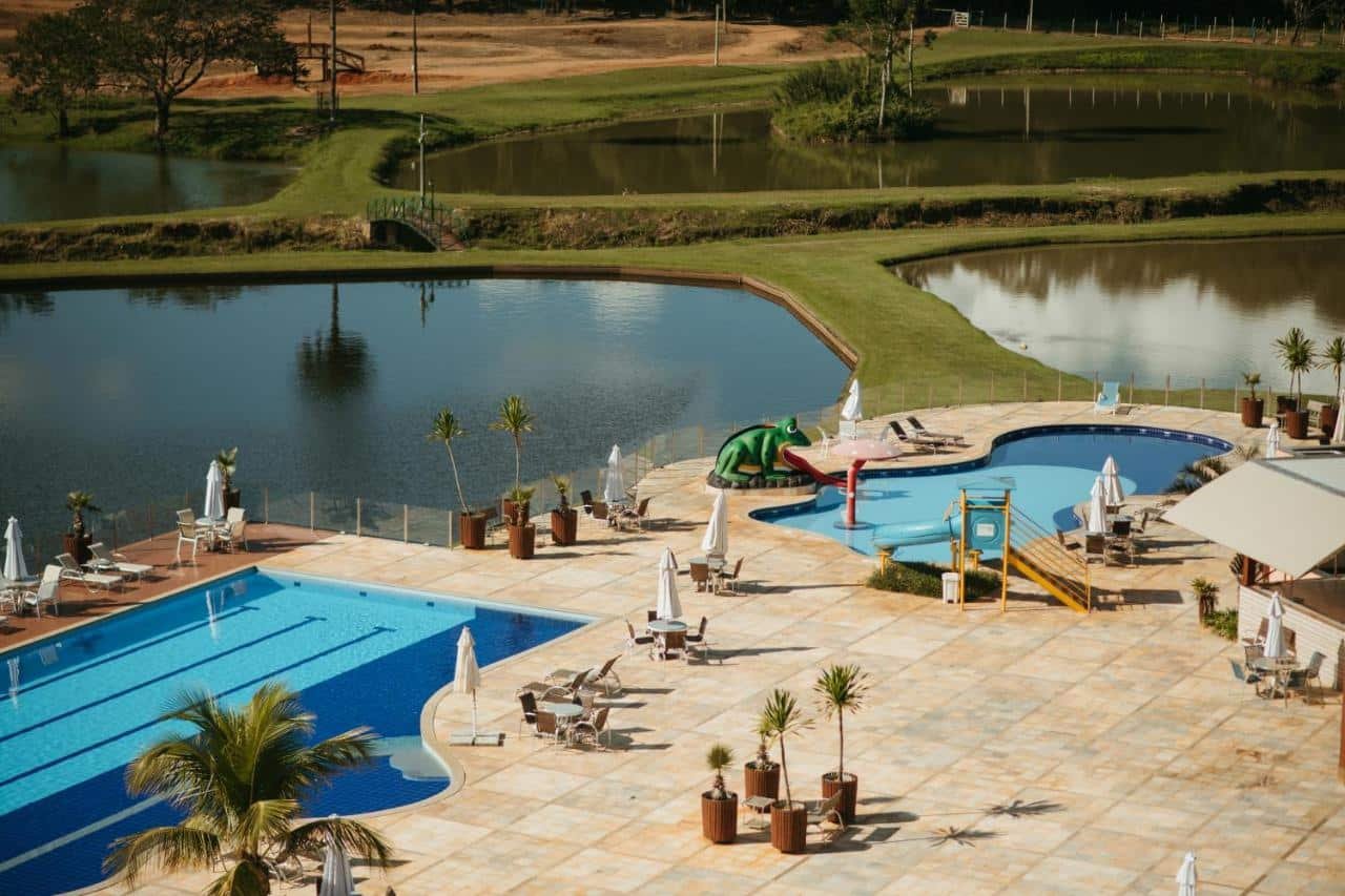 Vista aérea de uma área de piscina ao ar livre ao lado de um lago, com espreguiçadeiras, guarda-sóis e um pequeno toboágua. Esse local fica no Engenho da Serra Hotel EcoResort, um dos hotéis fazenda em Minas Gerais.