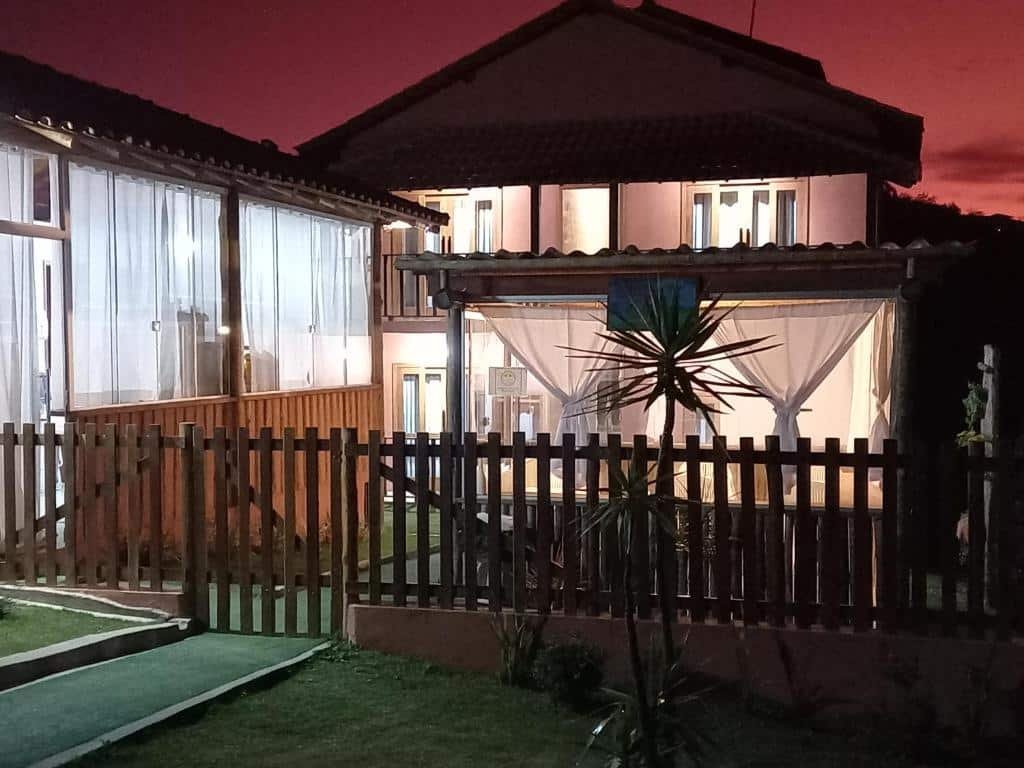 A imagem mostra a entrada da pousada Serra da Neblina em Lavras Novas, Minas Gerais, durante o entardecer. O céu está tingido de tons de rosa e roxo, indicando que o sol acabou de se pôr. A pousada possui uma construção rústica e charmosa, com detalhes em madeira. As janelas são amplas e estão fechadas com cortinas brancas, conferindo um ar acolhedor ao ambiente.
A entrada da pousada é cercada por uma cerca de madeira baixa, e há um caminho de grama bem cuidado que leva até a porta principal. À direita, pode-se ver uma área externa coberta com um pergolado e cortinas, criando um espaço aconchegante para relaxar. Plantas ornamentais estão espalhadas pelo jardim, complementando a decoração natural do local.
A iluminação interna da pousada está acesa, o que proporciona uma sensação de calor e hospitalidade.