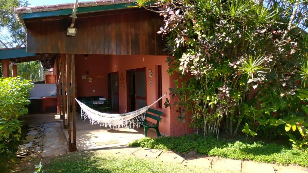 Entrada da Aconchego na Praia, uma casa pintada em vermelho com uma rede pendurada, muitas árvores e um jardim, além de um banco verde na varanda
