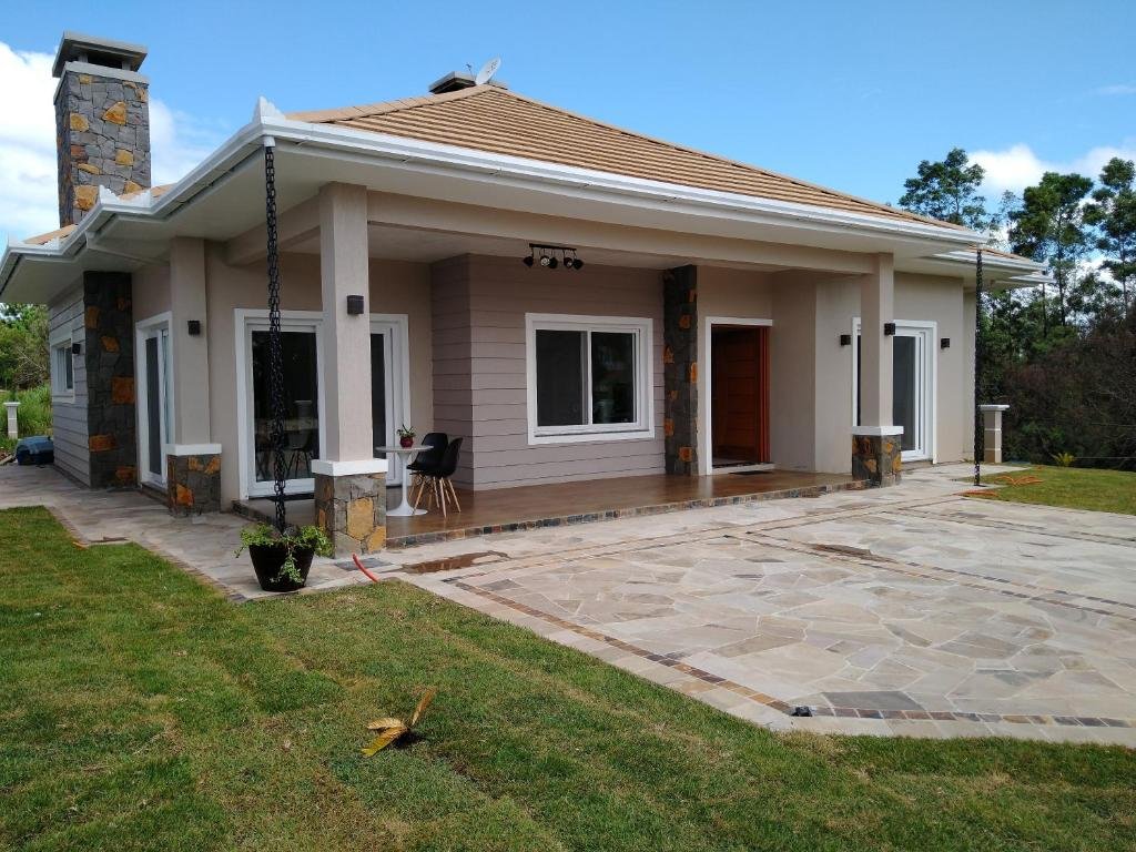 Fachada da Casa Gramado - Moradas da Serra, uma varanda com uma mesinha e duas cadeiras, um jardim ao redor, casa térrea e nova em tons de branco e cinza