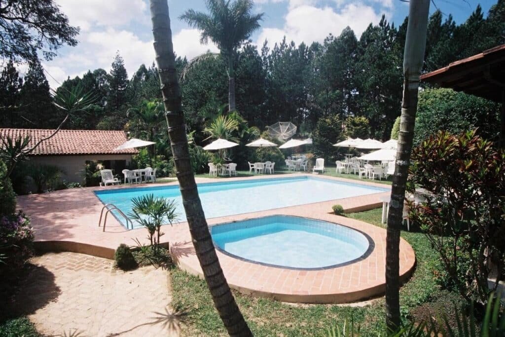 A imagem mostra uma piscina ao ar livre em uma área cercada por vegetação exuberante e palmeiras. Há duas piscinas, uma maior e uma menor, possivelmente destinada a crianças, ambas rodeadas por espreguiçadeiras e mesas com guarda-sóis brancos. O cenário é tranquilo, com muitas plantas ao redor e o céu azul parcialmente coberto por nuvens. Ao fundo, há uma construção com telhado de telhas vermelhas e cercada por árvores altas, criando um ambiente acolhedor e relaxante. Essa é a Estalagem Fazenda Lazer, um dos hotéis-fazenda em Minas Gerais.