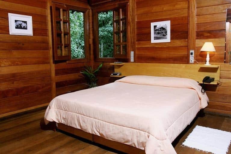 A imagem mostra um quarto aconchegante com paredes e piso de madeira, criando uma atmosfera rústica na Estalagem Fazenda Lazer. No centro do quarto, há uma cama de casal com lençóis claros e uma cabeceira de madeira. Ao lado da cama, há uma mesinha com um abajur aceso, proporcionando uma iluminação suave. As janelas de madeira estão abertas, revelando a vegetação verde do lado de fora. Na parede, há duas pequenas fotos emolduradas que complementam a decoração simples e acolhedora do ambiente.