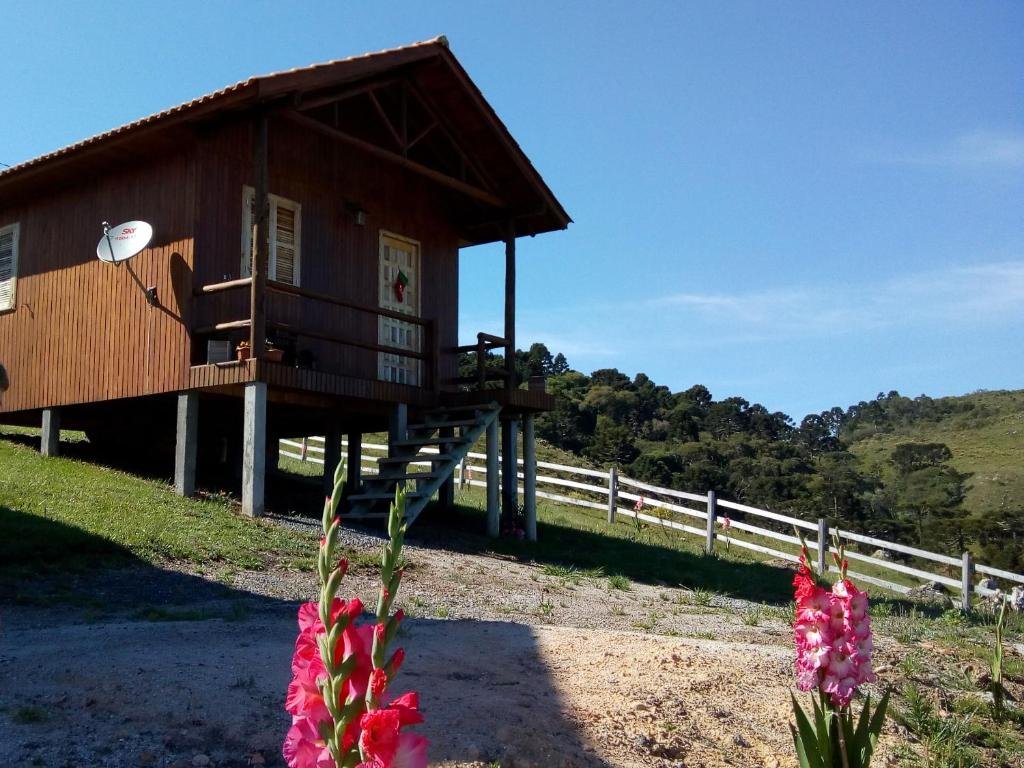 A imagem mostra uma cabana simples e elevada sobre pilares no Paraíso dos Cânions, um dos chalés na Serra Catarinense, localizada em uma área rural. A construção é de madeira, com um telhado inclinado e uma pequena varanda à frente, acessível por uma escada. Uma antena parabólica está fixada na lateral da cabana. Ao redor, vê-se um campo verde com cercas brancas e, ao fundo, colinas e vegetação densa. Algumas flores cor-de-rosa estão em primeiro plano, acrescentando cor ao cenário rural e tranquilo.