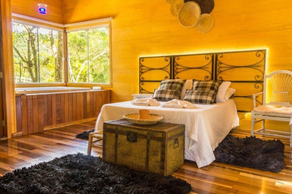 Um quarto aconchegante na Fazenda dos Caetés com interior de madeira, uma cama de casal, um baú antigo como mesa de cabeceira e uma grande janela com vista para a vegetação.
