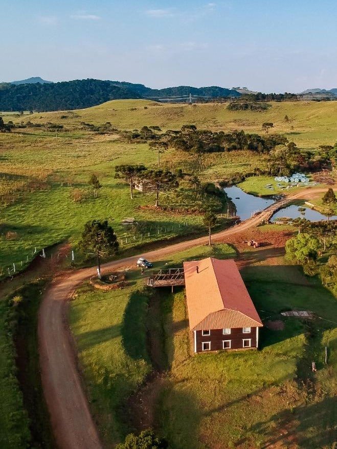 Vista aérea de uma paisagem rural com uma estrada de terra, uma casa com telhado vermelho, uma pequena ponte, árvores, um lago e colinas distantes na Fazenda Invernada Grande Turismo Rural, um dos hotéis fazenda em Santa Catarina.