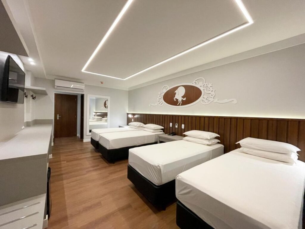 Um quarto do Fazzenda Park Resort com quatro camas de solteiro bem arrumadas, uma TV montada na parede, um espelho, uma escrivaninha, piso de madeira e decoração minimalista.